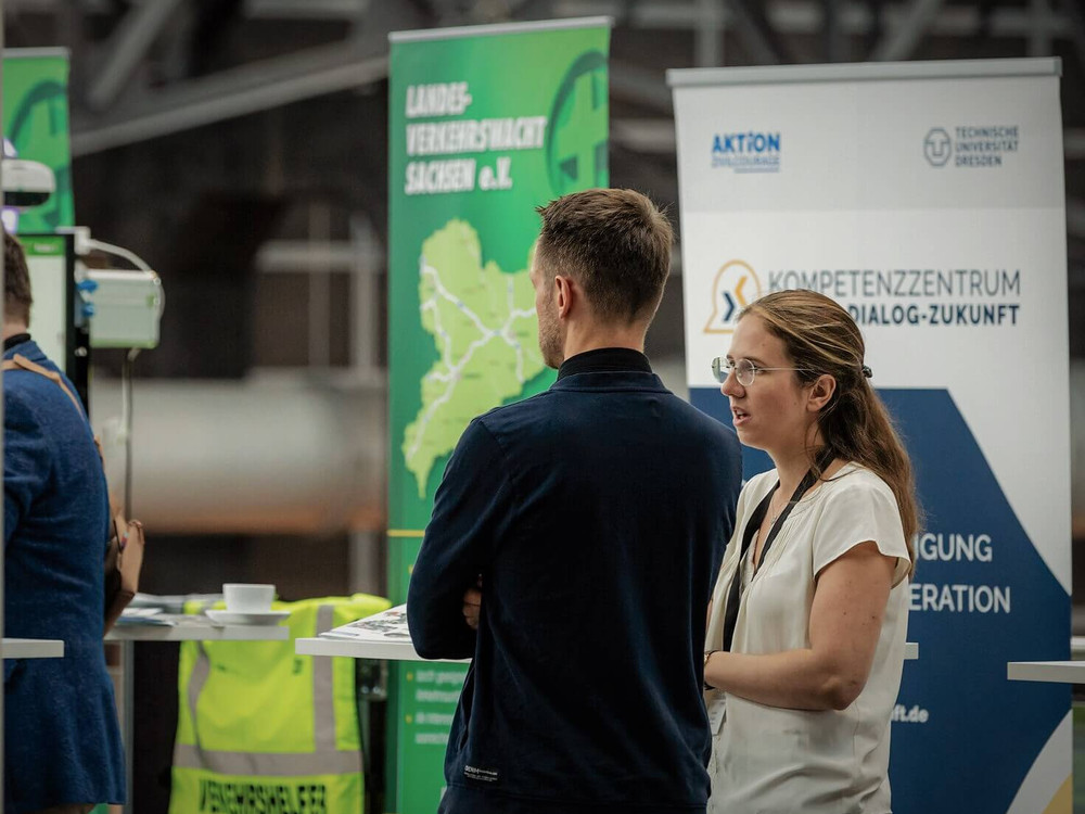 KI generiert: Das Bild zeigt zwei Personen, die sich auf einer Veranstaltung unterhalten, während im Hintergrund ein Stand mit einer Karte und dem Logo des "Kompetenzzentrum Dialog-Zukunft" zu sehen ist. Der Hauptinhalt des Bildes ist die Interaktion der Personen in einem professionellen oder informativen Umfeld.