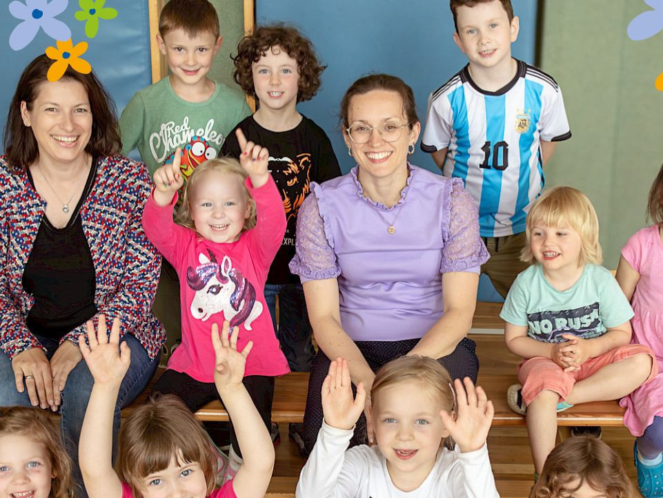 KI generiert: Das Bild zeigt eine Gruppe fröhlicher Kinder, die gemeinsam mit einer Erwachsenen in einem hellen Raum stehen und ihre Hände in die Luft strecken. Im Hintergrund sind zwei Puppen an der Fensterbank zu sehen.