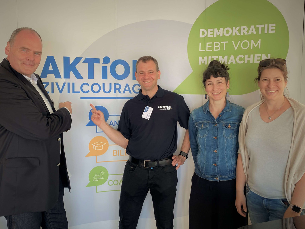 KI generiert: Das Bild zeigt einen Stand der "Aktion Zivilcourage" auf einer Veranstaltung, an dem zwei Personen stehen und Informationsmaterial anbieten. Im Hintergrund sind ein Plakat mit der Aufschrift "Demokratie lebt vom Mitmachen" und ein Glücksrad zu sehen.