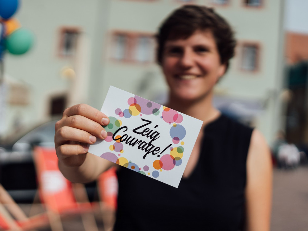 KI generiert: Das Bild zeigt eine lächelnde Person, die ein Kärtchen mit der Aufschrift "Zeig Courage!" in der Hand hält. Im Hintergrund sind bunte Ballons und eine Straßenumgebung zu erkennen.