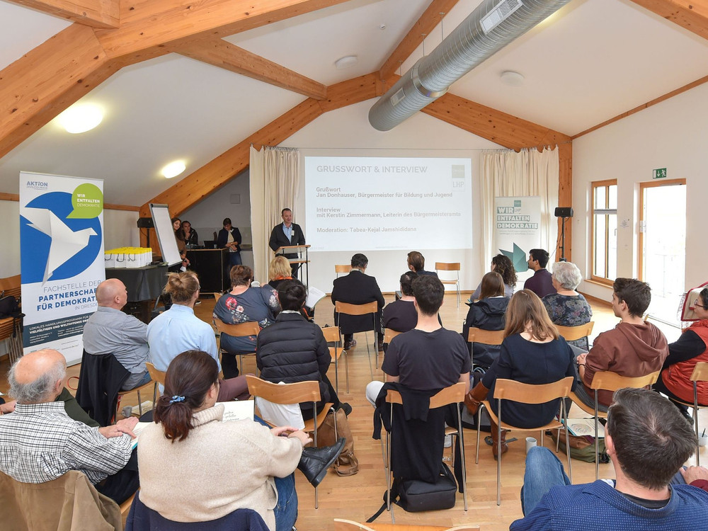KI generiert: Das Bild zeigt eine Versammlung oder Konferenz in einem hellen, holzvertäfelten Raum, bei der eine Person vorne steht und zu den Anwesenden spricht, während eine Präsentation auf einem Bildschirm im Hintergrund läuft. Der Hauptinhalt des Bildes ist eine formelle Präsentation oder Diskussion in einer Gruppenumgebung.