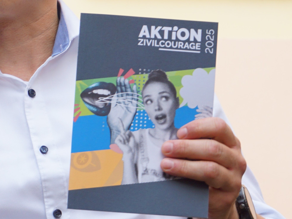 KI generiert: Das Bild zeigt eine Person, die ein Mikrofon in der Hand hält und ein Heft oder eine Broschüre mit dem Titel "AKTION" präsentiert. Im Hintergrund ist ein Fenster mit Gittern zu sehen.