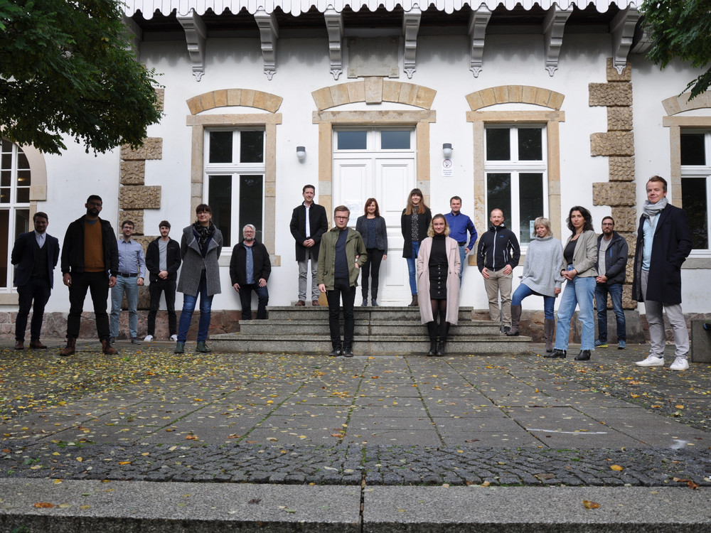 KI generiert: Das Bild zeigt eine Gruppe von 18 Personen, die vor einem Gebäude mit großen Fenstern und einer Treppe posieren. Die Personen sind in verschiedenen Kleidungsstilen gekleidet und scheinen sich für ein Gruppenfoto versammelt zu haben.