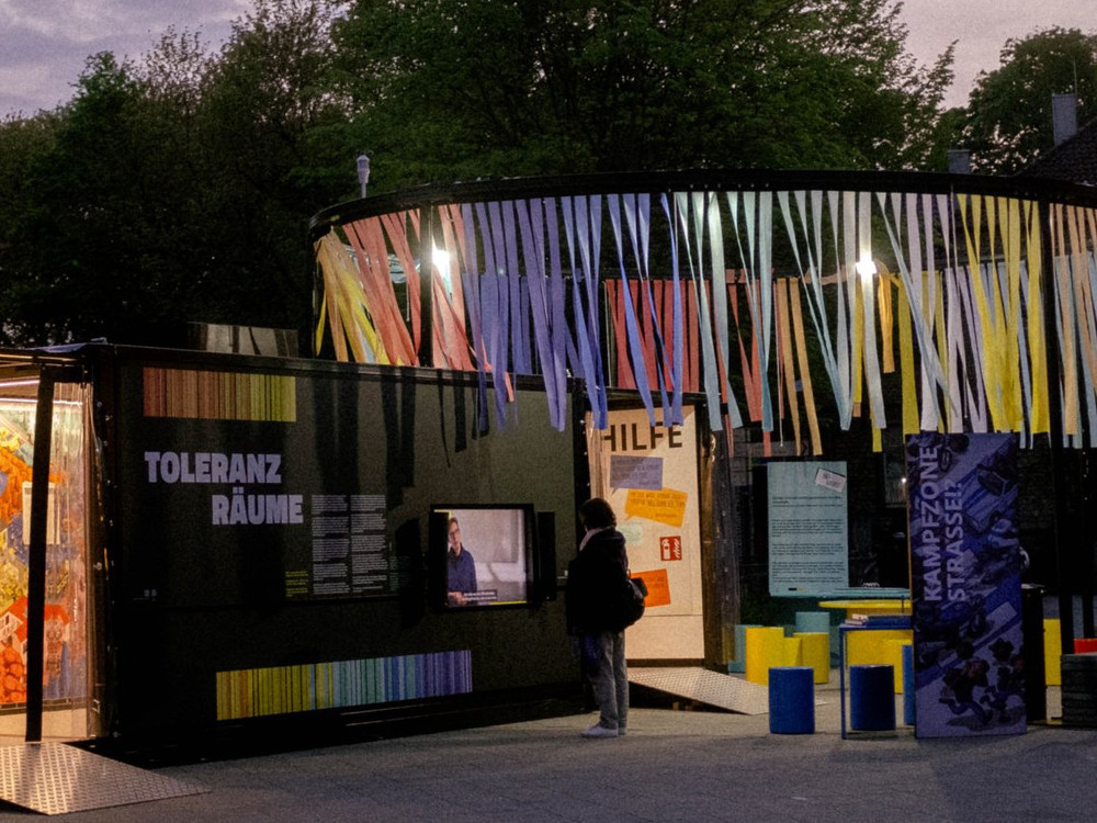 KI generiert: Das Bild zeigt eine belebte Szene mit Menschen, die sich um einen bunten Pavillon mit der Aufschrift "JOCKELS GASTANNAHME GANZ NAH" versammeln, während im Hintergrund ein historisches Gebäude und Bäume zu sehen sind. Der Hauptinhalt des Bildes ist der Pavillon und die Menschen, die sich dort aufhalten.