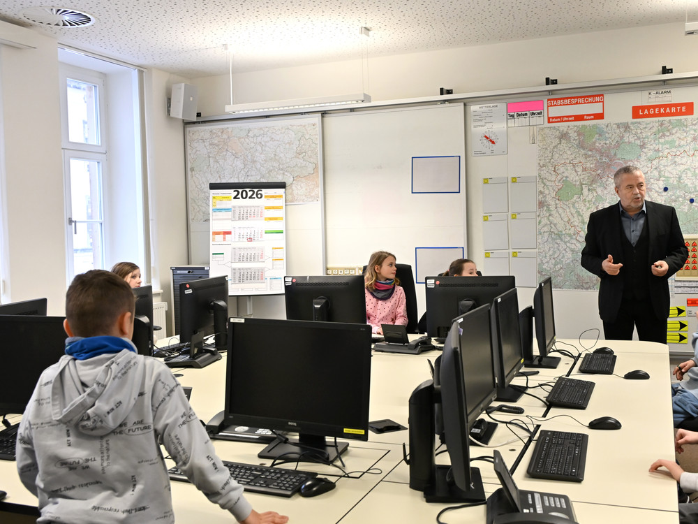 KI generiert: Das Bild zeigt einen Klassenraum, in dem ein Lehrer vor einer Gruppe von Schülern steht und unterrichtet. Die Schüler sitzen an Computern und scheinen einer Präsentation oder einem Vortrag zu folgen.