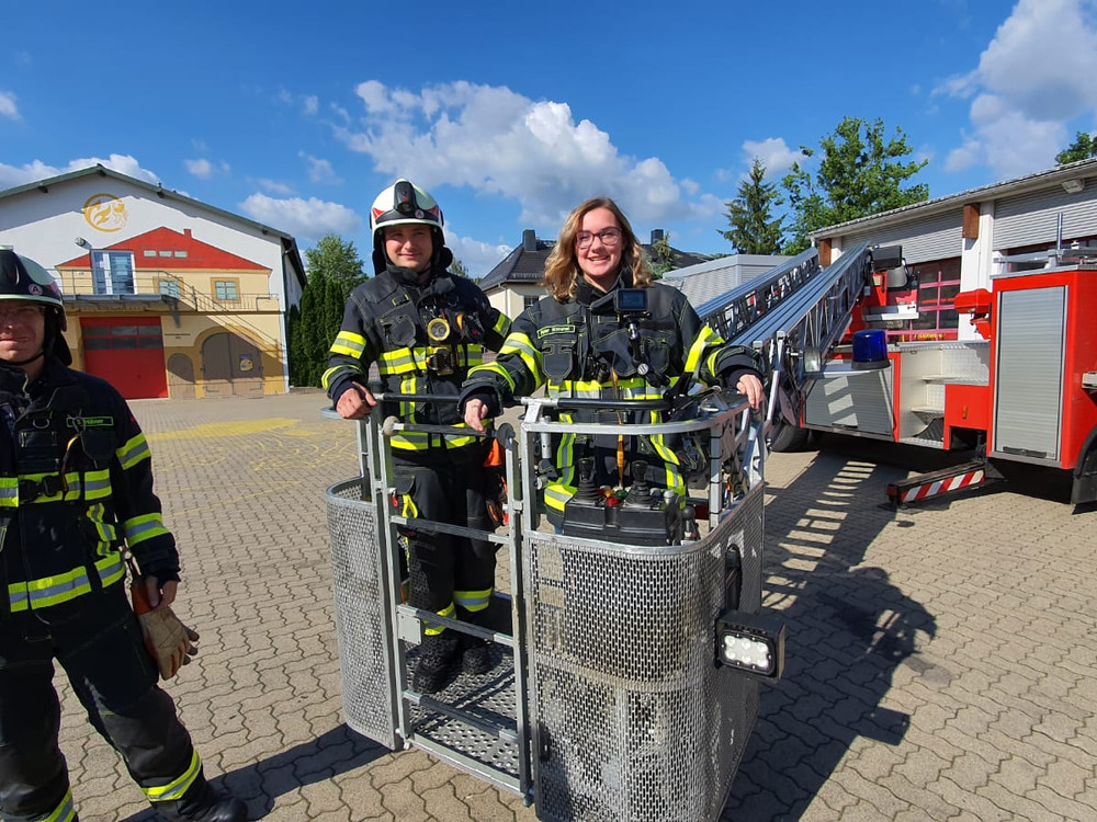 KI generiert: Das Bild zeigt drei Feuerwehrleute in voller Schutzausrüstung, die vor einem Feuerwehrfahrzeug und einer Feuerwache posieren. Im Vordergrund steht ein spezielles Rettungsgerät, möglicherweise eine Trage oder ein Transportbehälter.
