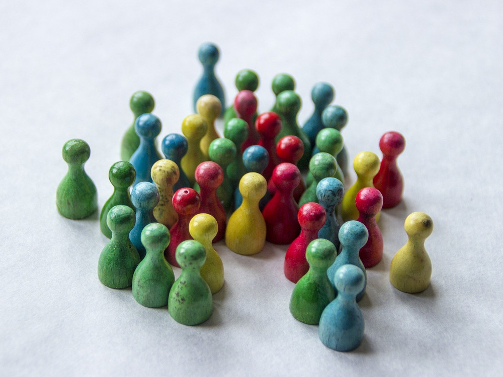 KI generiert: Das Bild zeigt eine kreisförmige Anordnung von bunt gezeichneten, stilisierten Menschenfiguren, die sich an den Händen halten und eine sonnenartige Formation bilden. Der Hauptinhalt des Bildes ist die Darstellung von Vielfalt und Gemeinschaft durch die verschiedenen Farben und die verbundenen Hände.