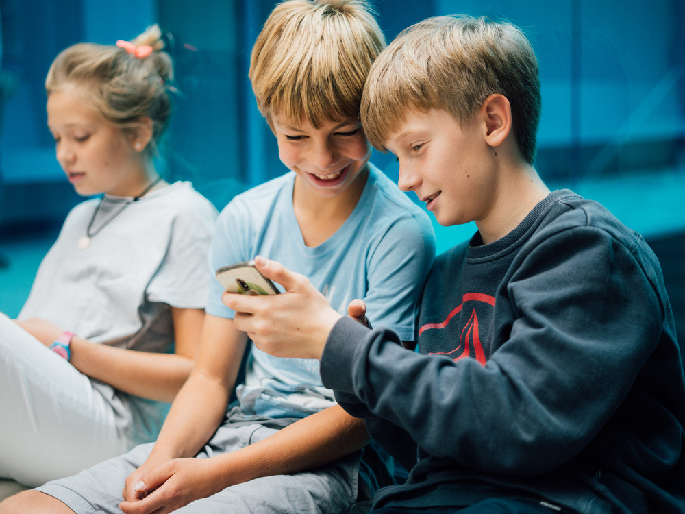 KI generiert: Das Bild zeigt drei Kinder, die eng beieinander sitzen und gemeinsam auf ein Smartphone schauen. Der Hauptinhalt des Bildes ist die Interaktion der Kinder mit dem Smartphone, was ihre Aufmerksamkeit und Neugier weckt.