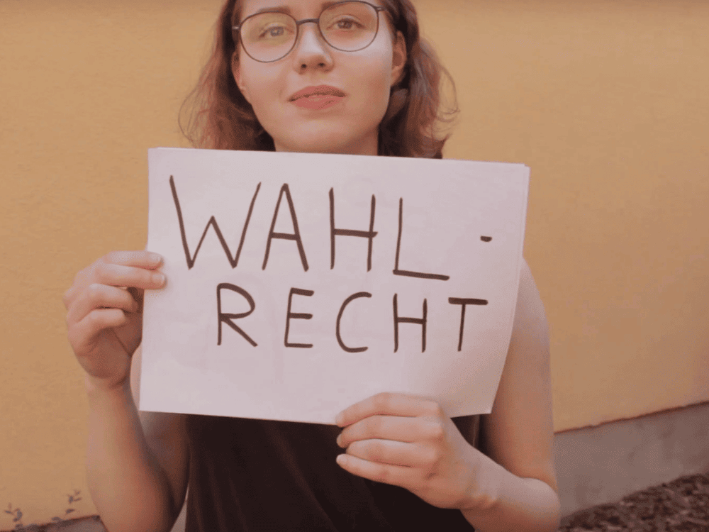 KI generiert: Das Bild zeigt eine Person, die ein weißes Blatt Papier mit der Aufschrift "WAHL-RECHT" in die Kamera hält. Der Hauptinhalt des Bildes ist die Forderung nach Wahlrecht.