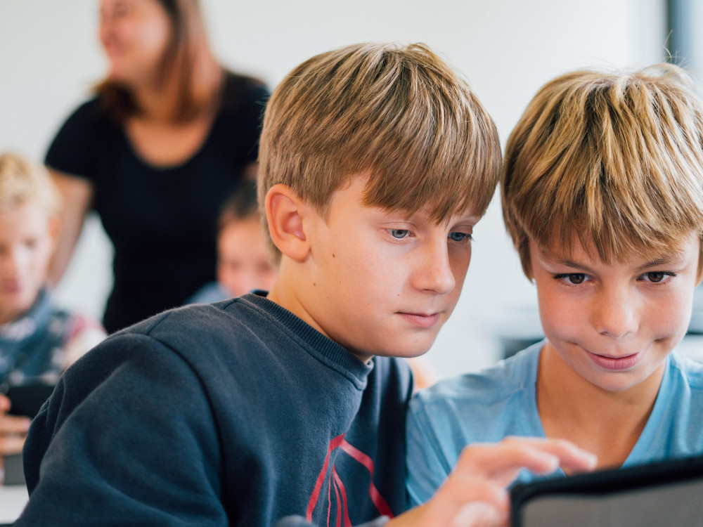 KI generiert: Das Bild zeigt zwei Jungen, die gemeinsam auf ein Tablet schauen, während sie in einem Klassenzimmer sitzen. Im Hintergrund sind weitere Kinder und eine erwachsene Person zu erkennen.