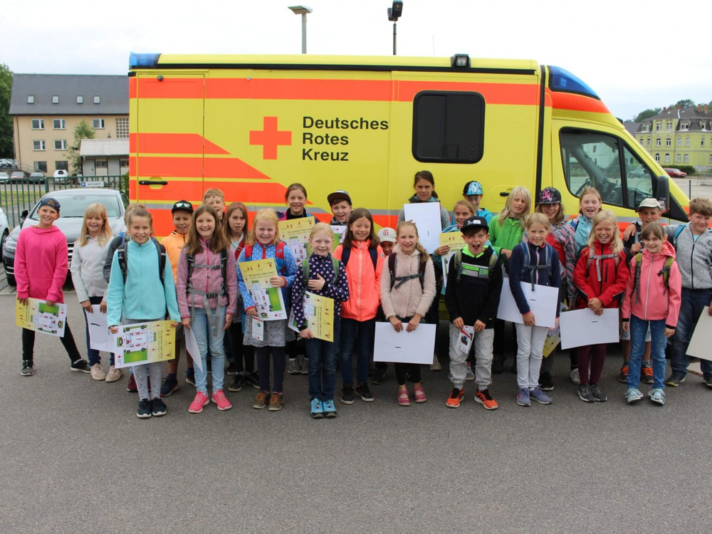 KI generiert: Das Bild zeigt eine Gruppe von Kindern, die vor einem Rettungswagen des Deutschen Roten Kreuzes (DRK) stehen und stolz ihre Zertifikate oder Urkunden präsentieren. Der Hauptinhalt des Bildes ist die feierliche Übergabe von Auszeichnungen, möglicherweise im Rahmen einer Erste-Hilfe-Ausbildung oder eines ähnlichen Programms.