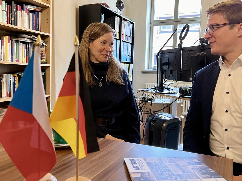 KI generiert: Das Bild zeigt eine Gruppe von sechs Personen, die vor einem Banner mit der Aufschrift "AKTION Zivilcourage" und den Flaggen von Tschechien und Deutschland stehen. Der Hauptinhalt des Bildes ist die Darstellung einer Zusammenarbeit oder eines Treffens im Rahmen einer Initiative für Zivilcourage.
