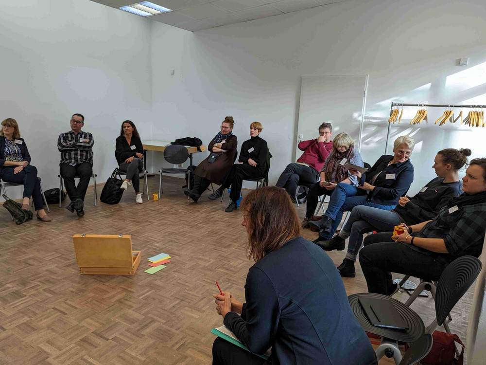 KI generiert: Das Bild zeigt eine Gruppe von Menschen, die in einem hellen, geräumigen Raum mit hohen Decken und großen Fenstern stehen und sich unterhalten. Im Raum gibt es mehrere Stehtische mit weißen Tischdecken, an denen einige Personen stehen, sowie eine kleine Bühne oder ein Podium im Hintergrund.