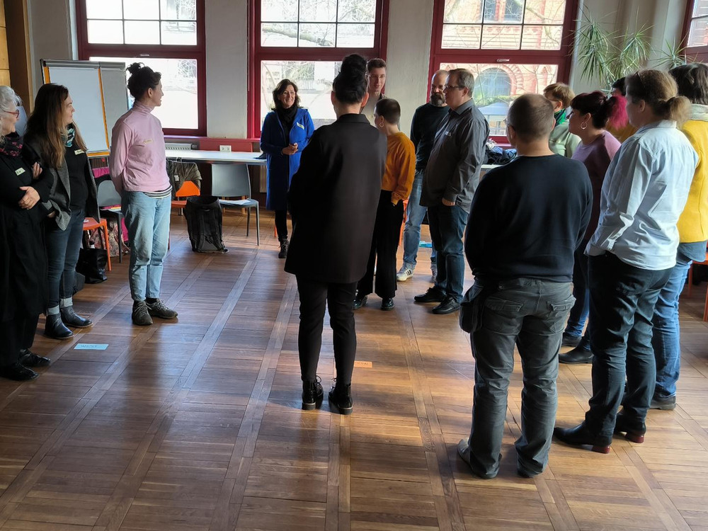 KI generiert: Das Bild zeigt eine Gruppe von Menschen, die in einem hellen Raum mit großen Fenstern stehen und sich unterhalten. Der Hauptinhalt des Bildes ist eine informelle Zusammenkunft oder ein Treffen, bei dem die Teilnehmer in kleinen Gruppen diskutieren.