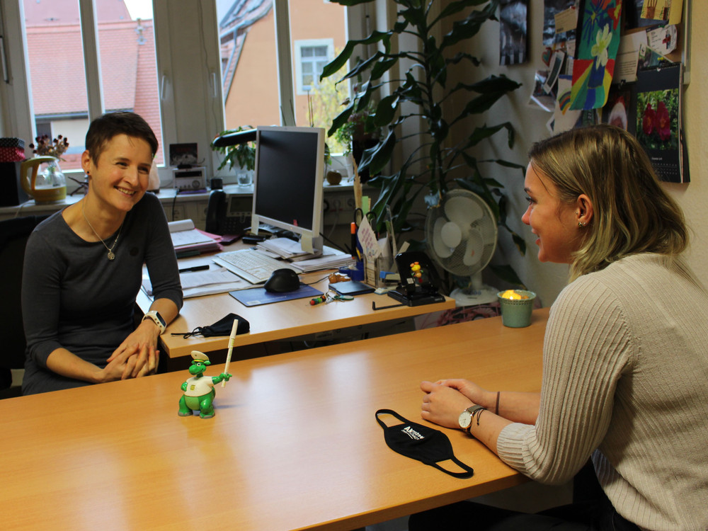 KI generiert: Das Bild zeigt zwei Personen in einem Büro, wobei eine Person an einem Schreibtisch sitzt und die andere Person steht und mit ihr spricht. Der Hauptinhalt des Bildes ist eine Büroumgebung mit Computern, Pflanzen und verschiedenen Büromaterialien.