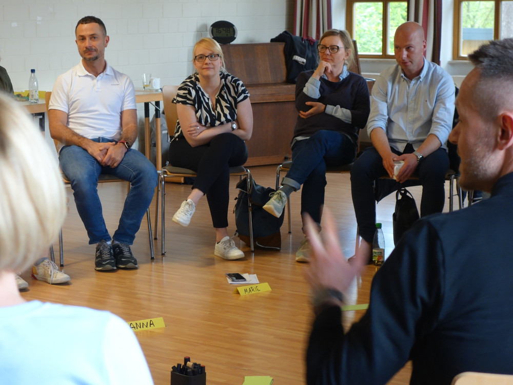 KI generiert: Das Bild zeigt eine Gruppe von Menschen in einem Seminarraum, wobei einige auf Stühlen im Kreis sitzen und eine Person vorne steht und präsentiert. Der Hauptinhalt des Bildes ist eine Diskussion oder ein Workshop in einer Bildungs- oder Schulungseinrichtung.