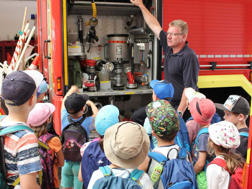 KI generiert: Das Bild zeigt eine Gruppe von Kindern, die neugierig die offene Tür eines Feuerwehrautos umringen, während ein Mann in schwarzem Shirt ihnen etwas erklärt. Der Hauptinhalt des Bildes ist eine Bildungsveranstaltung, bei der Kinder die Ausrüstung eines Feuerwehrautos kennenlernen.