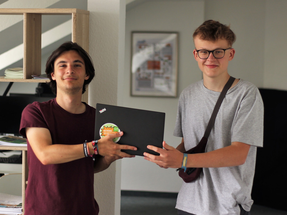 KI generiert: Das Bild zeigt zwei junge Männer, die vor Laptops sitzen und in die Kamera lächeln. Der Hauptinhalt des Bildes ist eine Arbeits- oder Lernumgebung, in der die beiden Personen möglicherweise zusammenarbeiten oder studieren.