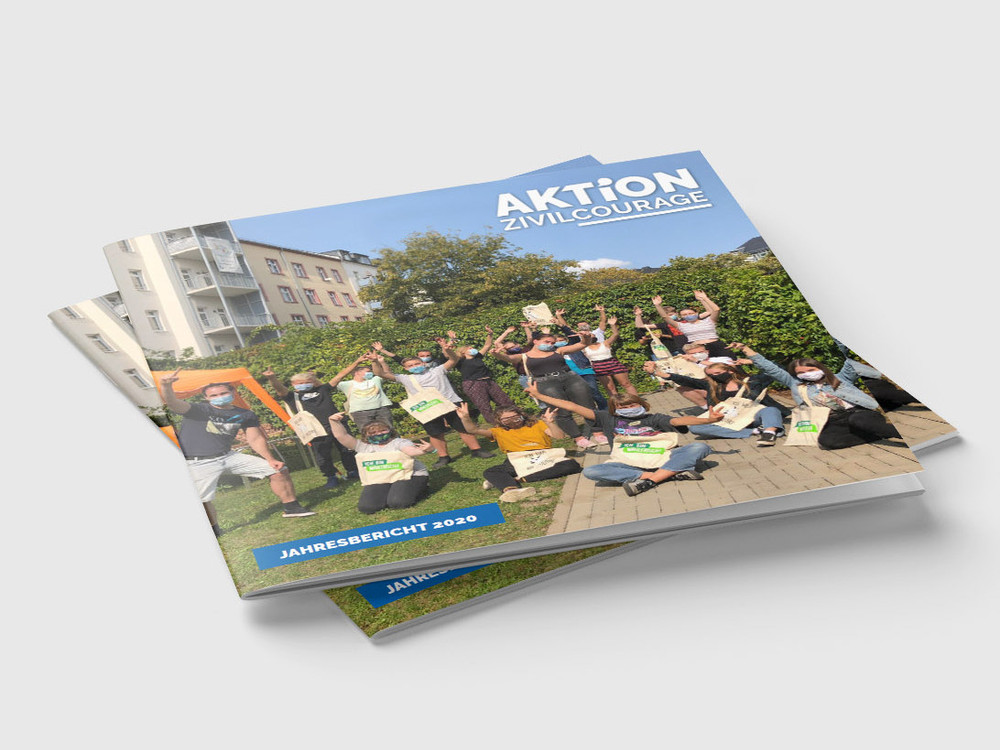 KI generiert: Das Bild zeigt mehrere Exemplare eines Jahresberichts mit dem Titel "Aktion Zivilcourage" auf einem Stapel. Der Hauptinhalt des Bildes ist der Jahresbericht selbst, der auf dem Cover eine Gruppe von Menschen in einer entspannten, geselligen Atmosphäre im Freien zeigt.