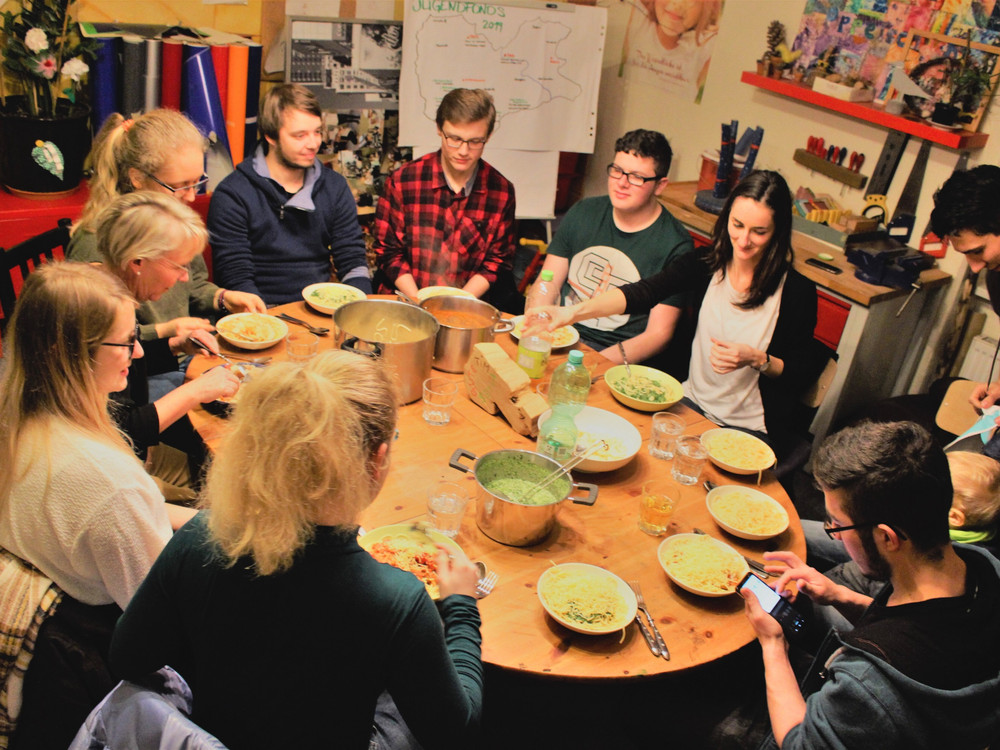 KI generiert: Das Bild zeigt eine Gruppe von Menschen, die gemeinsam an einem großen Holztisch sitzen und verschiedene Gerichte aus großen Töpfen servieren. Der Hauptinhalt des Bildes ist ein geselliges Essen in einer gemütlichen, informellen Atmosphäre.