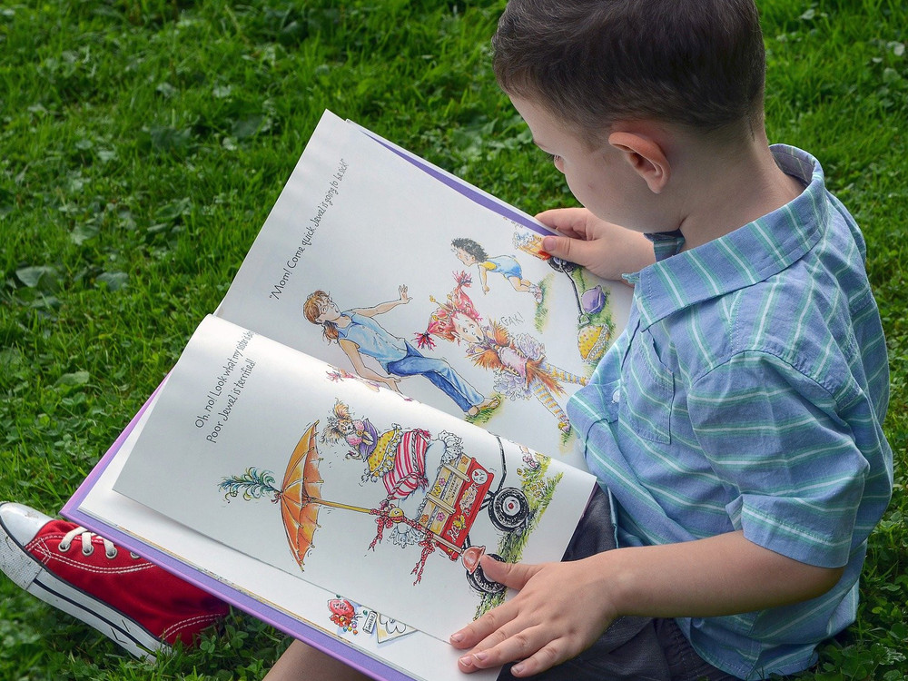 KI generiert: Das Bild zeigt einen Jungen, der auf dem Rasen sitzt und ein buntes Kinderbuch liest. Der Hauptinhalt des Bildes ist das Lesen und die kindliche Freude am Entdecken von Geschichten.