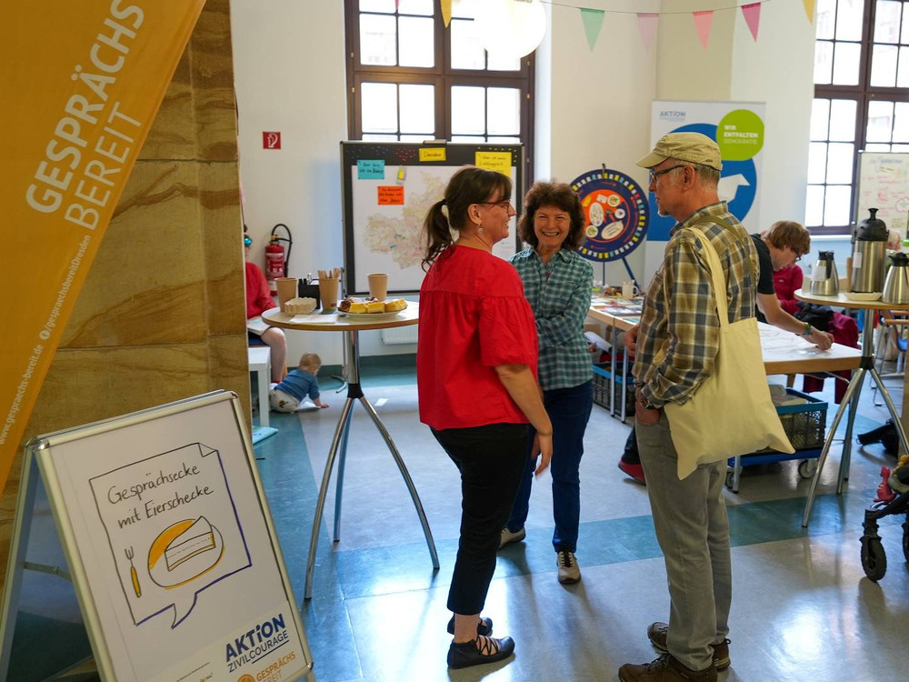 KI generiert: Das Bild zeigt eine Ausstellungs- oder Informationsveranstaltung in einem hellen, historischen Raum mit mehreren Ständen und Postern. Der Hauptinhalt ist der "DemokratieMarktPlatz", wie auf dem Banner über den Ständen zu lesen ist.
