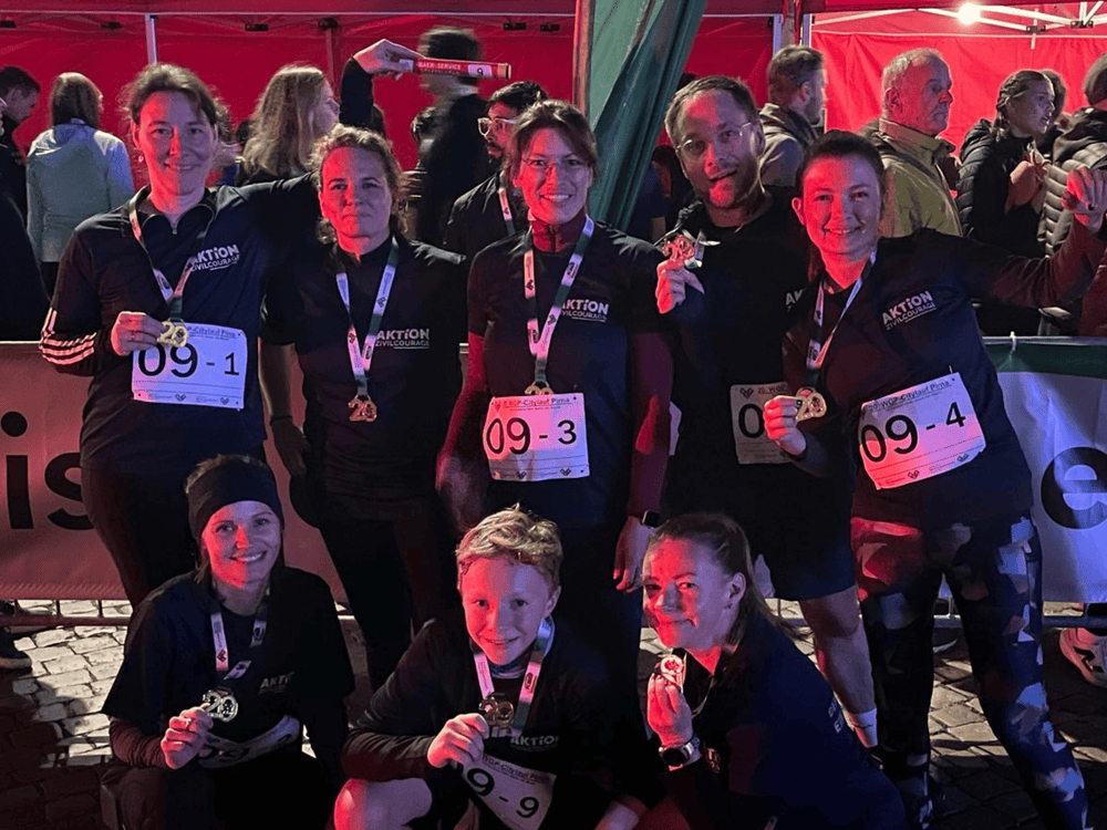 KI generiert: Das Bild zeigt eine Gruppe von Personen, die an einem Event teilnehmen, möglicherweise einem Sportwettbewerb oder einer Gemeinschaftsveranstaltung. Die Teilnehmer tragen Nummernschilder und Medaillen, was darauf hindeutet, dass sie eine Aktivität abgeschlossen haben.