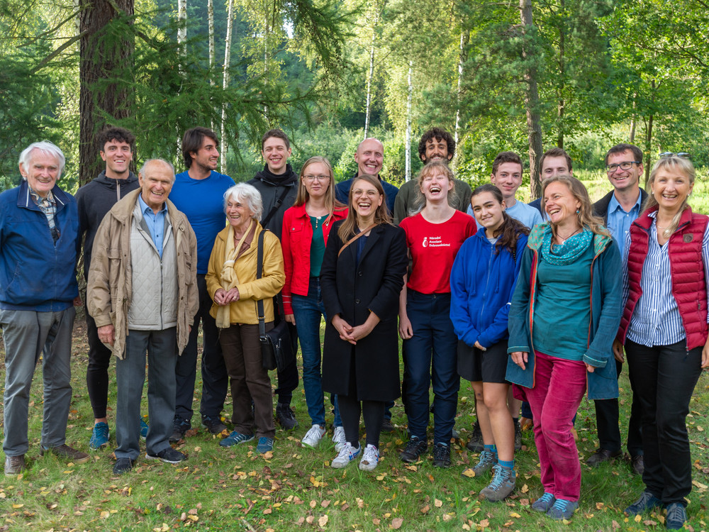 KI generiert: Das Bild zeigt eine Gruppe von Menschen, die in einer natürlichen Umgebung, möglicherweise einem Wald oder Park, zusammenstehen und lächeln. Der Hauptinhalt des Bildes ist die Darstellung einer fröhlichen und vielfältigen Gruppe in einer Outdoor-Umgebung.