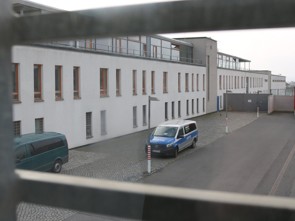 KI generiert: Das Bild zeigt einen Innenhof mit gepflastertem Boden, umgeben von Gebäuden mit weißen Fassaden und mehreren Fenstern. Im Vordergrund steht ein blauer Polizeiwagen, und im Hintergrund sind ein roter LKW sowie weitere Fahrzeuge zu sehen.