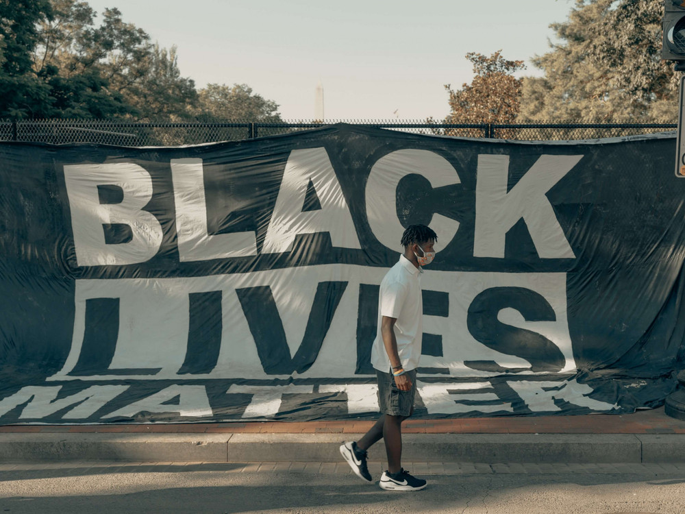 KI generiert: Das Bild zeigt eine Person, die an einem großen Banner mit der Aufschrift "BLACK LIVES MATTER" vorbeigeht. Das Banner ist an einem Zaun befestigt, und im Hintergrund sind Bäume und ein Verkehrsschild zu sehen.