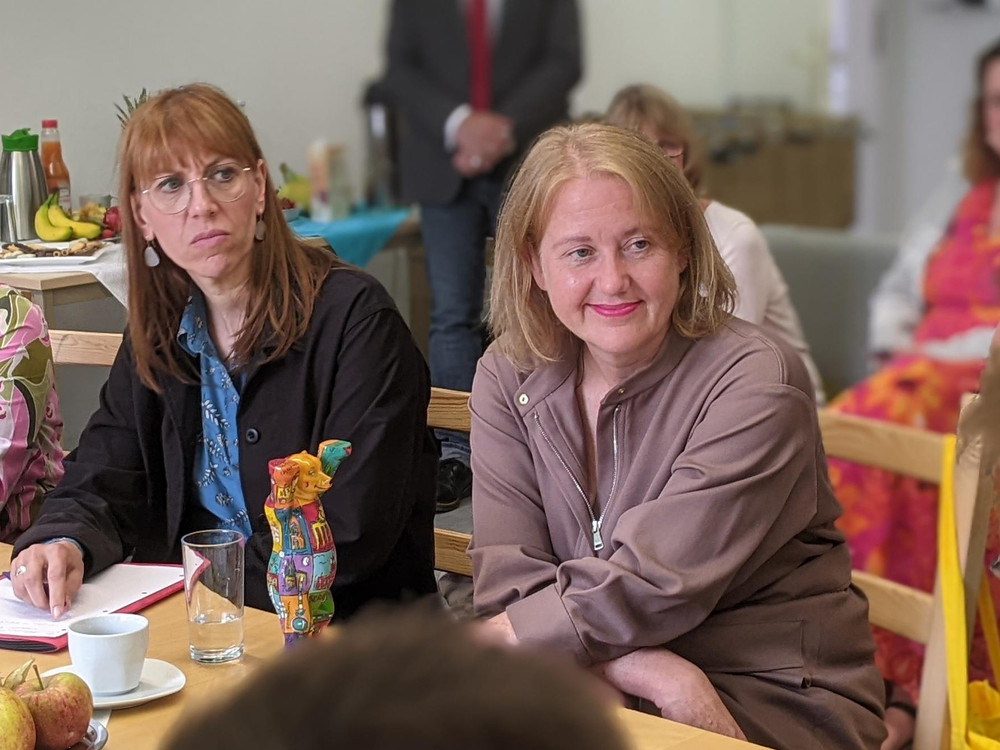 KI generiert: Das Bild zeigt eine Gruppe von Personen, die sich im Freien auf einem gepflasterten Platz unterhalten. Im Vordergrund stehen zwei Personen im Gespräch, während im Hintergrund weitere Menschen in kleinen Gruppen zusammenstehen und sich unterhalten.
