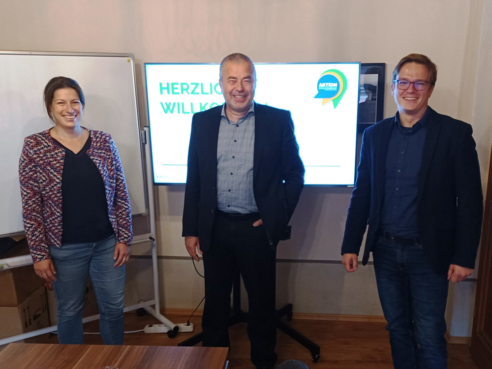 KI generiert: Das Bild zeigt drei lächelnde Personen, die vor einem Plakat mit der Aufschrift "HERZLICH WILLKOMMEN" und dem Logo "AKTION" stehen. Der Hauptinhalt des Bildes ist eine Begrüßungsszene, möglicherweise bei einer Veranstaltung oder einem Meeting.