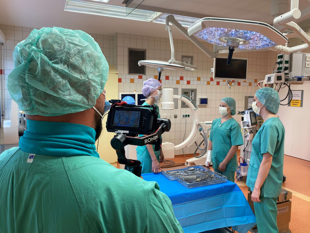 KI generiert: Das Bild zeigt eine Gruppe von medizinischem Personal in einem Operationssaal, die sich auf eine Operation vorbereiten. Im Vordergrund ist eine Person mit einer Kameraausrüstung zu sehen, die möglicherweise die Operation aufzeichnet.