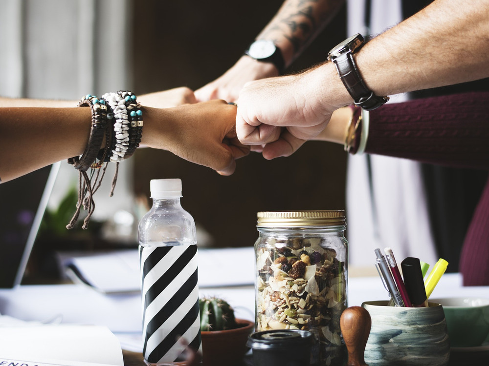 KI generiert: Das Bild zeigt mehrere Hände, die sich in der Mitte zu einem festen Handschlag vereinen, was Teamwork und Zusammenhalt symbolisiert. Die Personen tragen verschiedene Armbänder und Uhren, was auf Individualität hinweist.