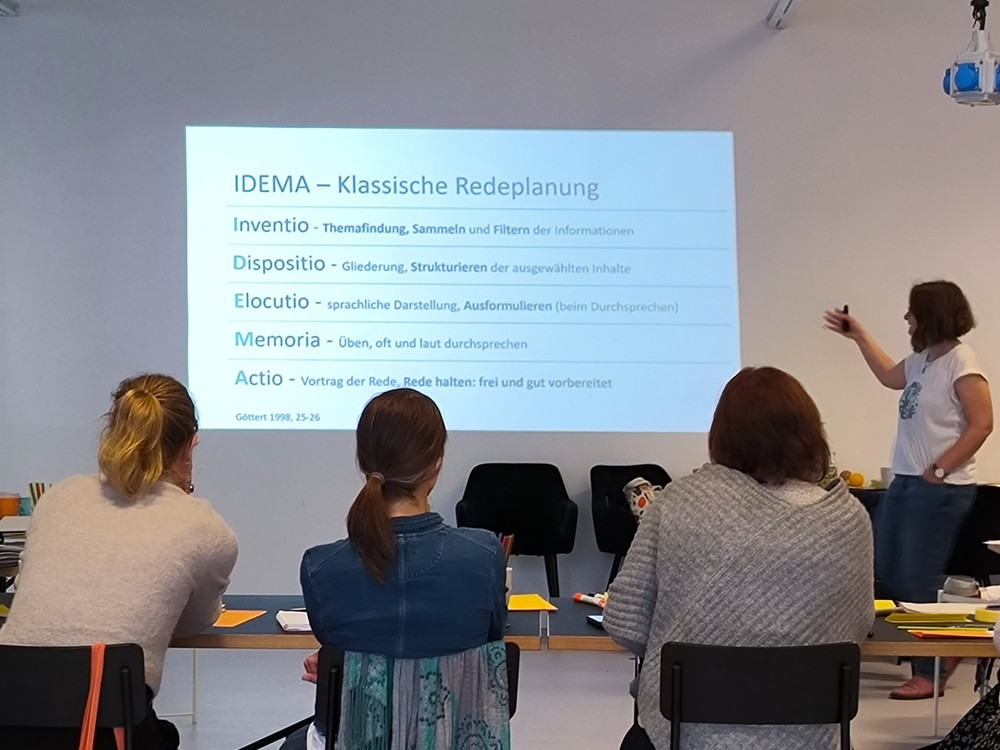 KI generiert: Das Bild zeigt eine Person, die vor einer Gruppe von Zuhörern steht und eine Präsentation hält. Der Hauptinhalt der Präsentation auf der Leinwand ist die klassische Redeplanung nach der IDEMA-Methode, die die Schritte Inventio, Dispositio, Elocutio, Memoria und Actio umfasst.