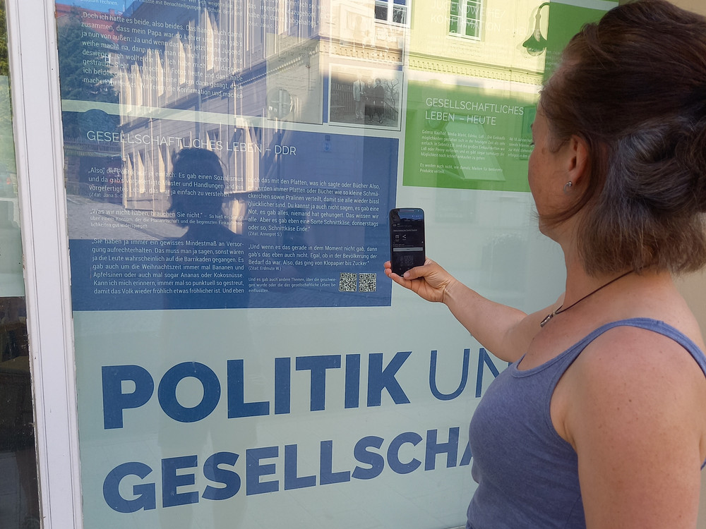 KI generiert: Das Bild zeigt eine Person, die mit einem Smartphone einen QR-Code scannt, der auf einem Plakat an einer Glaswand zu sehen ist. Der Hauptinhalt des Bildes ist das Scannen des QR-Codes, der wahrscheinlich zu weiteren Informationen über das Thema des Plakats führt.