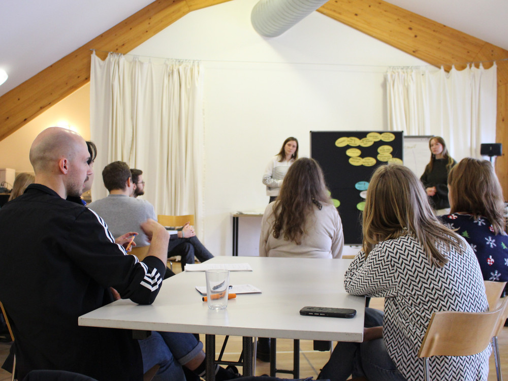 KI generiert: Das Bild zeigt eine Gruppe von sechs Personen, die auf Stühlen in einer Reihe sitzen und sich in einer lockeren Runde unterhalten. Der Hauptinhalt des Bildes ist eine informelle Diskussion oder ein Workshop in einem Innenraum.