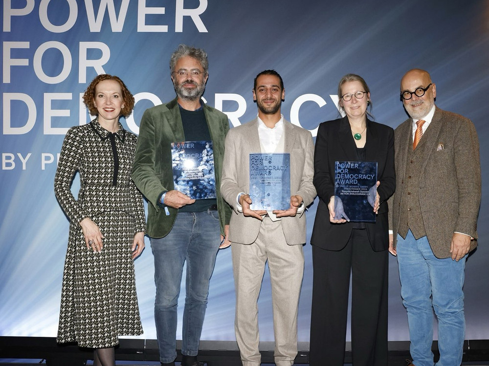 KI generiert: Das Bild zeigt fünf Personen, die auf einer Bühne stehen und jeweils eine Urkunde oder einen Preis in den Händen halten. Der Hintergrund deutet auf eine Veranstaltung hin, bei der ein "Democracy Award" verliehen wird.