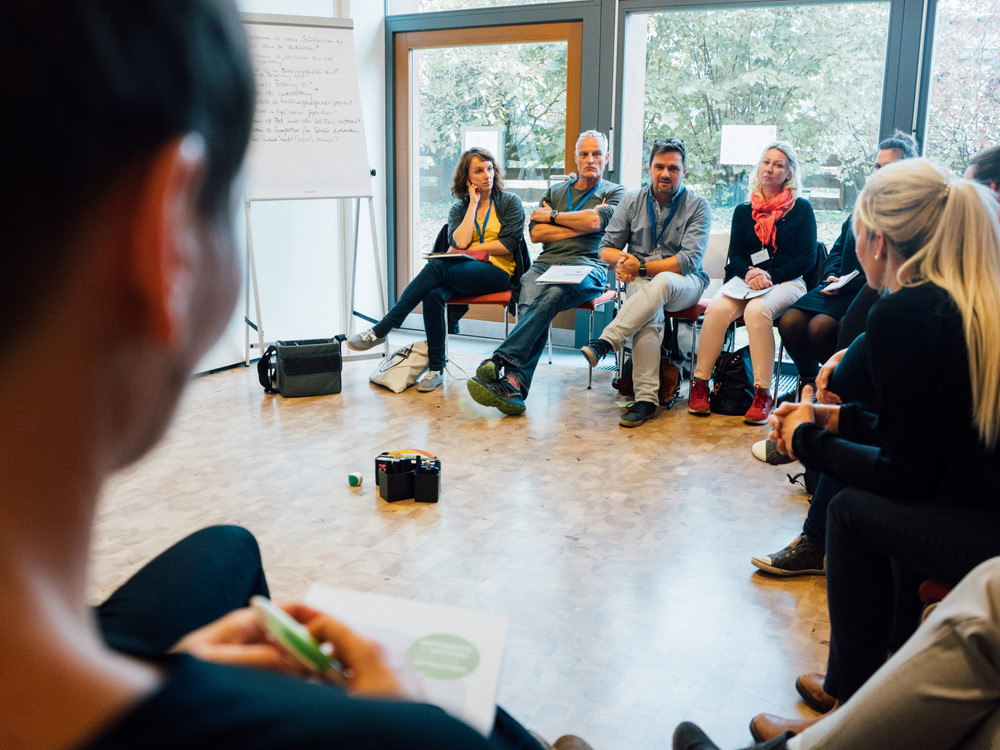 KI generiert: Das Bild zeigt eine Gruppe von Personen, die in einem Kreis sitzen und sich in einer Besprechung oder einem Workshop befinden. Der Hauptinhalt des Bildes ist eine interaktive Sitzung oder Diskussion in einem professionellen oder pädagogischen Umfeld.