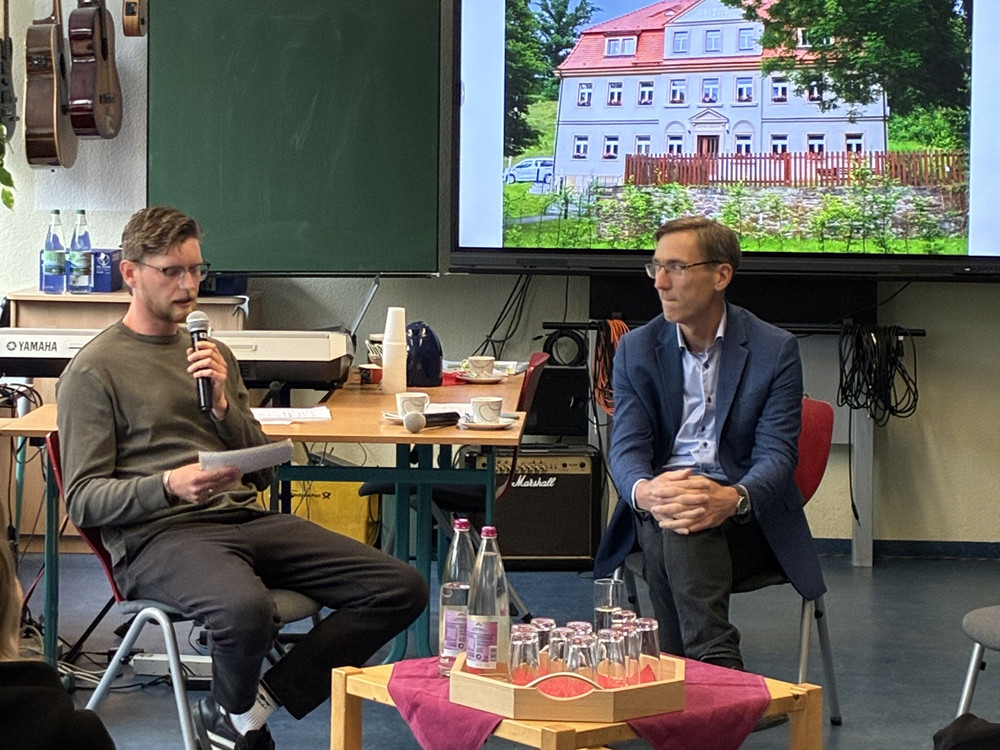 KI generiert: Das Bild zeigt eine Gruppe von vier Personen, die in einem Klassenzimmer an Tischen sitzen und sich unterhalten. Im Hintergrund sind eine Tafel, Musikinstrumente und Pflanzen zu sehen, was auf eine entspannte Diskussion oder ein Meeting in einer schulischen oder kreativen Umgebung hindeutet.