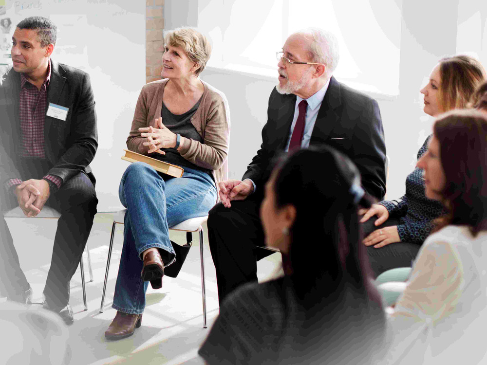 KI generiert: Das Bild zeigt eine Gruppe von Personen in einem Seminarraum, wobei ein Mann vorne steht und zu den Teilnehmern spricht. Im Hintergrund sitzt eine Frau an einem Tisch mit einem Mikrofon, während die Zuhörer auf Stühlen Platz genommen haben.
