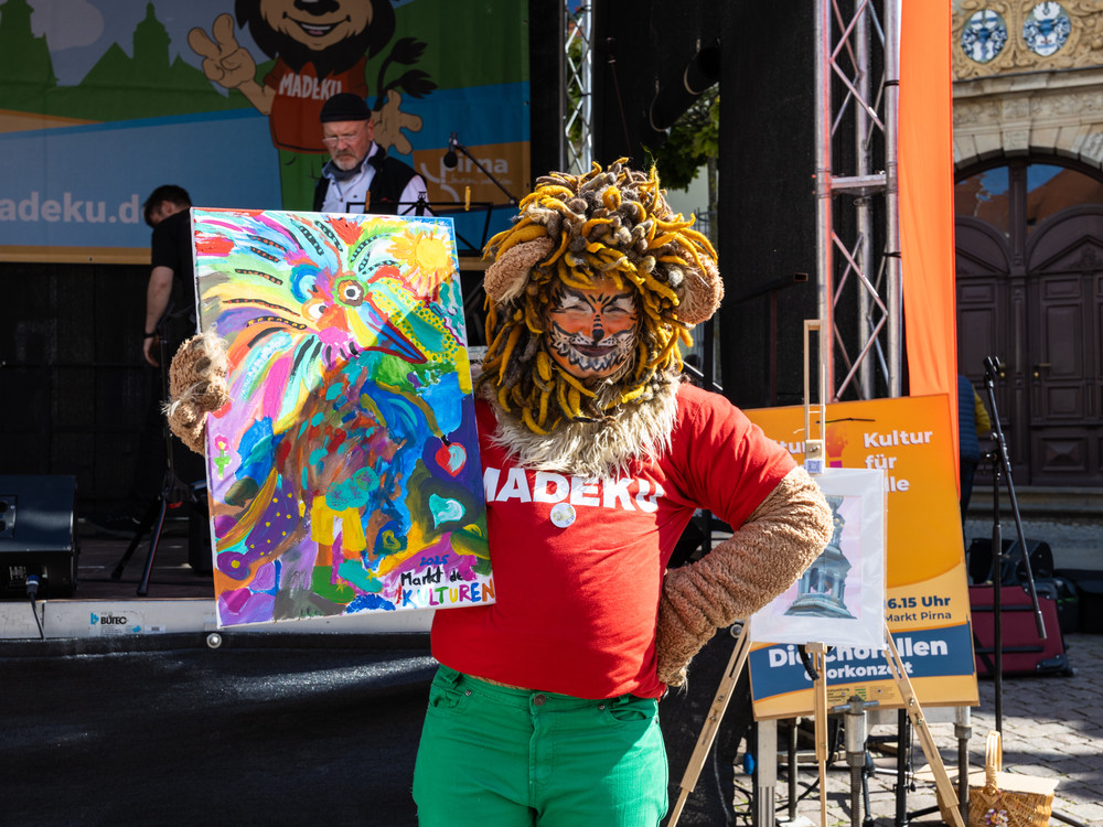 KI generiert: Das Bild zeigt eine belebte Open-Air-Veranstaltung auf einem Platz in einer Stadt mit historischen Gebäuden, bei der eine Band auf einer Bühne auftritt und viele Menschen zuschauen. Der Hauptinhalt des Bildes ist das kulturelle Event mit Live-Musik und einem großen Publikum.