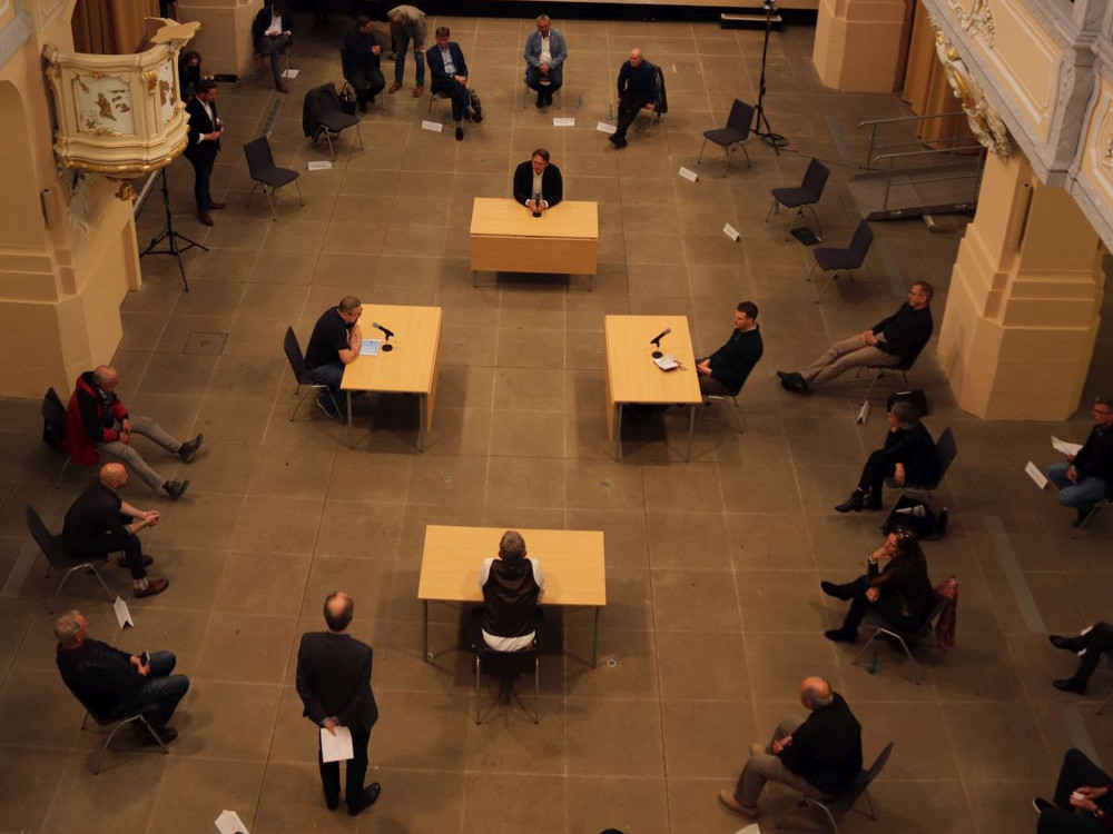 KI generiert: Das Bild zeigt eine Gruppe von Personen, die in einem großen Raum an mehreren runden Tischen sitzen und sich unterhalten. Der Hauptinhalt des Bildes ist eine Versammlung oder ein Treffen, bei dem die Teilnehmer in kleinen Gruppen diskutieren.