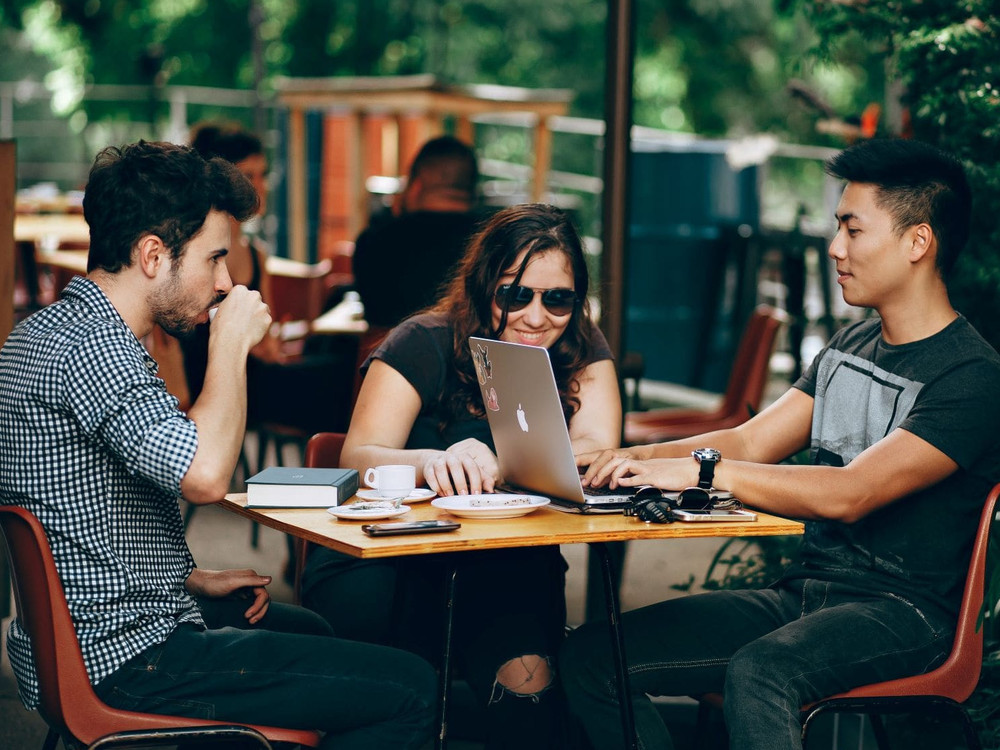 KI generiert: Das Bild zeigt drei Personen, die an einem Tisch sitzen und gemeinsam an einem Laptop arbeiten, vermutlich in einem Café oder einer informellen Arbeitsumgebung. Der Hauptinhalt des Bildes ist die Zusammenarbeit und Diskussion der Gruppe.
