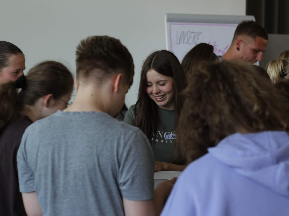 KI generiert: Das Bild zeigt eine Gruppe von Personen, die auf dem Boden sitzen und stehend in einem Innenraum posieren, wobei viele von ihnen mit den Händen winken. Der Hauptinhalt des Bildes ist eine fröhliche und entspannte Gruppensituation, möglicherweise eine Schulklasse oder eine Gruppe von Teilnehmern einer Veranstaltung.
