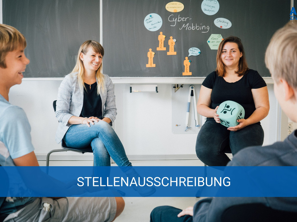 KI generiert: Das Bild zeigt eine Gruppe von Personen, darunter zwei Frauen und zwei Jugendliche, die in einem Klassenzimmer sitzen und sich unterhalten. Im Hintergrund ist eine Tafel mit Notizen und Zeichnungen zum Thema "Cyber-Mobbing" zu sehen. Der Hauptinhalt des Bildes ist eine Diskussion oder ein Workshop zum Thema Cyber-Mobbing.