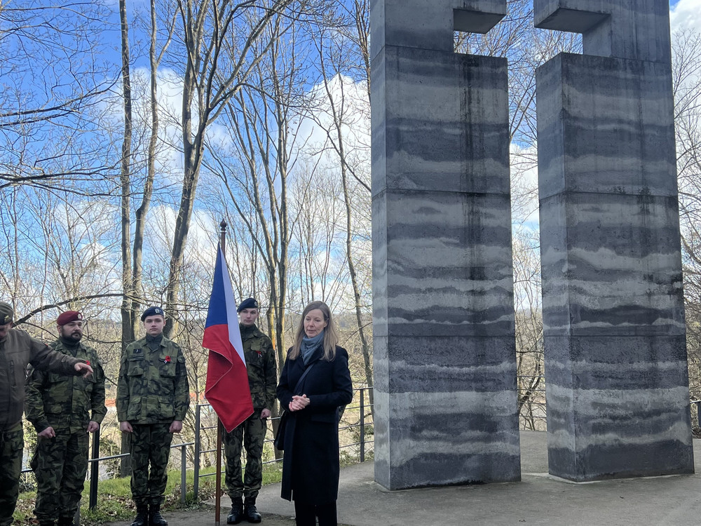 KI generiert: Das Bild zeigt eine Gruppe von Soldaten in Uniform, die an einer Gedenkveranstaltung teilnehmen, wobei eine Frau eine Rede vor einem Denkmal hält. Im Hintergrund ist eine Flagge zu sehen, die zwischen den Bäumen weht.
