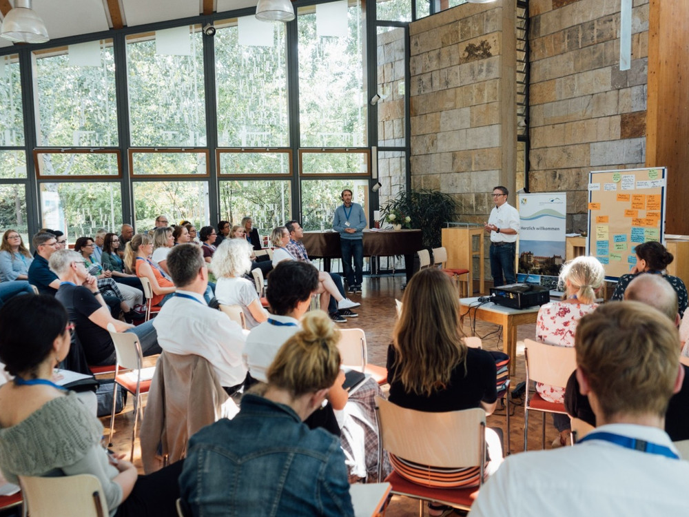 KI generiert: Das Bild zeigt eine Gruppe von Menschen, die in einem hellen, modernen Raum mit großen Fenstern an einer Präsentation oder einem Seminar teilnehmen. Der Hauptinhalt ist eine Versammlung oder ein Workshop, bei dem ein Mann vorne steht und zu den Anwesenden spricht.