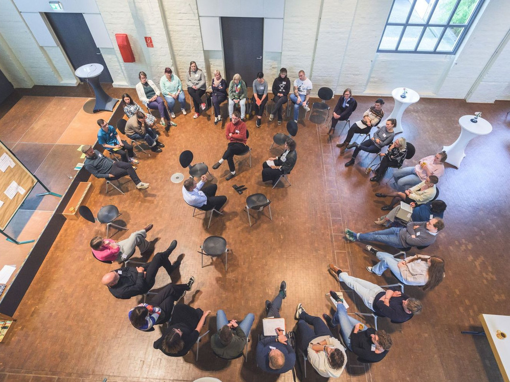 KI generiert: Das Bild zeigt eine Gruppe von Menschen, die in einem Kreis sitzen und sich in einer informellen Besprechung oder Diskussion befinden. Der Hauptinhalt des Bildes ist eine interaktive Sitzung oder ein Workshop in einem hellen, modernen Innenraum.