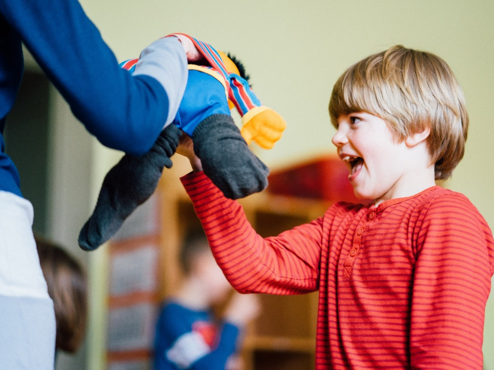 KI generiert: Das Bild zeigt ein Kind in einem roten gestreiften Oberteil, das freudig einen bunten Stoffesel von einer erwachsenen Person entgegennimmt. Im Hintergrund sind weitere Kinder in einer hellen, kindgerechten Umgebung zu erkennen.